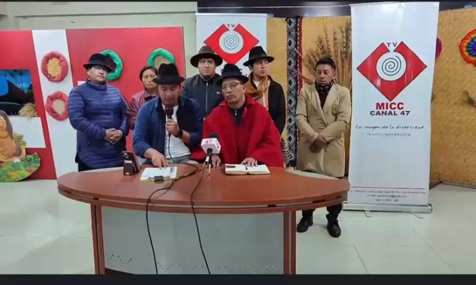 Several individuals, including a person in a red coat seated at a round wooden table with microphones, are gathered in a studio. Behind them, banners display the logo and name "MICC Canal 47" with a circular design featuring a hat. The setting includes red panels with wreaths and a mural of a child, alongside beige partitions with grain imagery. The individuals, some wearing hats and coats, stand or sit, with one holding a microphone and another holding documents. Text overlays are visible on the banners and panels.