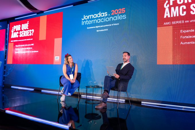 Blue-lit stage with promotional banners reading Jornadas Internacionales 2025, AMC Series expansion, and Por que AMC, featuring woman with brown hair in blue outfit and lanyard sitting on chair with legs crossed holding notebook, and man in dark suit with laptop on table, glass of water visible, black floor reflecting lights.