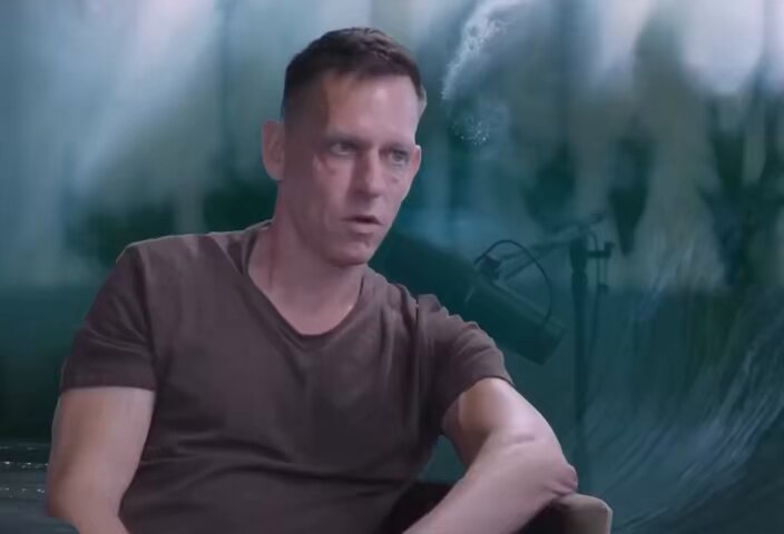 A man in a brown t-shirt sitting in front of a large, dynamic wave background, symbolizing the turbulent nature of the crypto market.