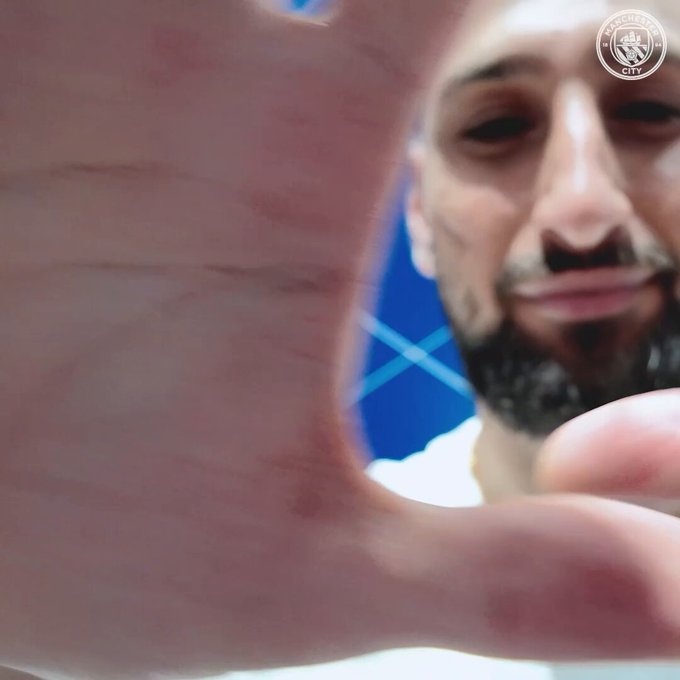 A video sequence showing a welcome message for Gianluigi Donnarumma to Manchester City. The first frame displays a close-up of a pink fabric with the Manchester City logo in the top right corner. The second frame features a man with a beard, wearing a white shirt, standing in front of a blue background with white geometric lines, saying 