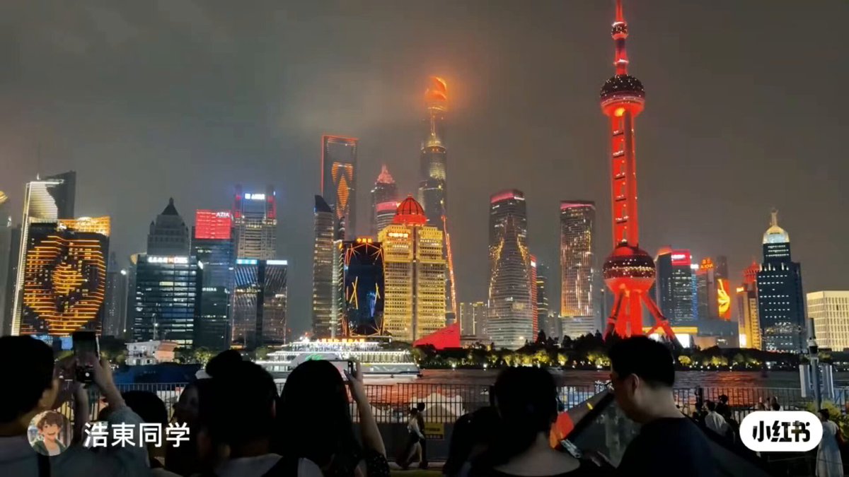 A nighttime panoramic view of Shanghai's Pudong skyline across the Huangpu River, featuring the illuminated Oriental Pearl Tower in red and gold, surrounded by modern skyscrapers with colorful LED lights and architectural highlights; in the foreground, a crowd of people, including tourists, takes photos from the Bund waterfront area with a boat visible on the river.