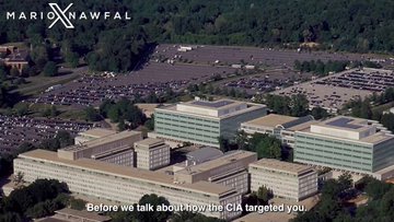 Aerial overhead view of a large government complex with multiple low-rise beige buildings surrounded by green trees, parking lots with cars, and roads; the facility appears secure with no visible people, set in a wooded suburban area.