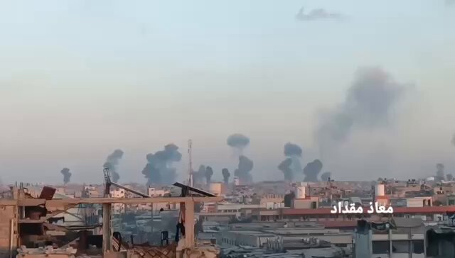 Aerial or elevated view of an urban area with multiple plumes of black smoke rising from various points amid a cityscape of low-rise buildings, some damaged structures, palm trees, and a hazy sky at dusk or dawn; no people or vehicles are clearly visible, focusing on the smoke and skyline.