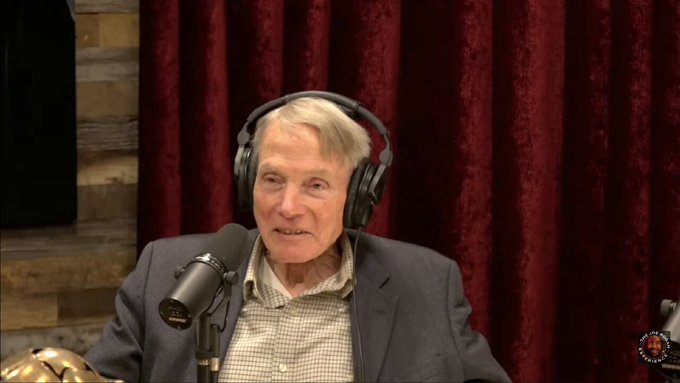 A screenshot from a podcast video showing an elderly white man with white hair, wearing a dark suit, light blue checkered shirt, and black headphones, speaking into a microphone while seated in a studio with wooden panel walls and red velvet curtains behind him. A small circular inset in the bottom right corner features a close-up of a man with a beard (Joe Rogan) wearing headphones.