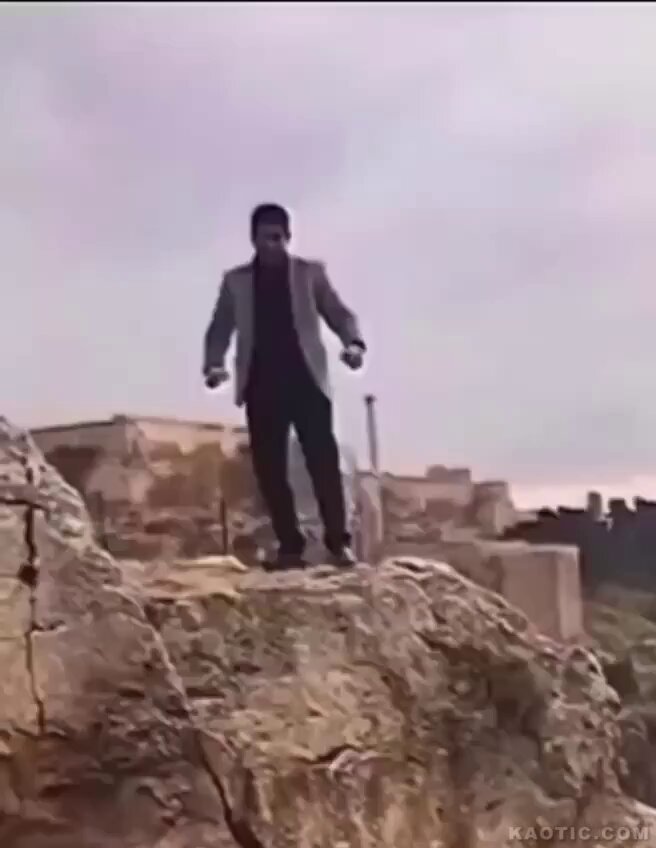 A man wearing a dark suit and white shirt stands on the edge of a rocky or concrete ledge with arms outstretched as if preparing to jump, against a cloudy sky; the background includes urban structures like buildings and pipes, suggesting a rooftop or artificial edge rather than a natural cliff; the image appears to be a frame from a video with a watermark in the bottom right.