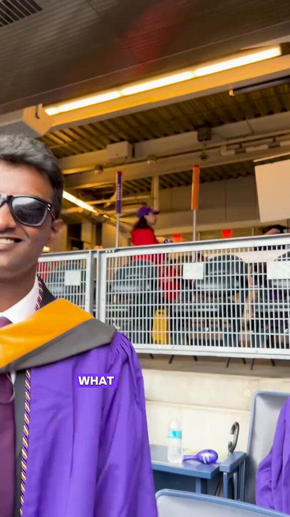 The image shows a smiling man of apparent South Asian descent wearing sunglasses and a purple graduation gown with orange and gray accents, standing in what appears to be an indoor stadium or arena during a commencement ceremony; in the background, there are bleachers, other graduates in similar gowns, a woman in a purple cap, and event signage or barriers.