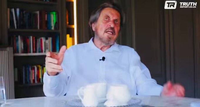 A frame from a video interview showing an elderly white man with gray hair and a beard, wearing a light blue shirt and a lapel microphone, gesturing emphatically with his hands while seated at a white table with two white teacups and a glass of water; behind him is a modern bookshelf filled with books and a wooden panel wall with warm lighting.