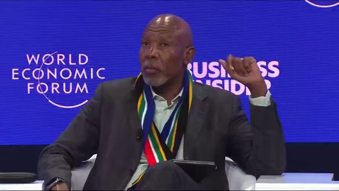 A middle-aged Black man with a bald head and graying beard, wearing a dark suit, light blue shirt, and a scarf in South African flag colors (black, green, yellow, white, red, blue), sits on a stage gesturing with his right hand while speaking into a microphone. The background features a large blue screen with the World Economic Forum logo and 'Business Insider' text, indicating a panel discussion setting.