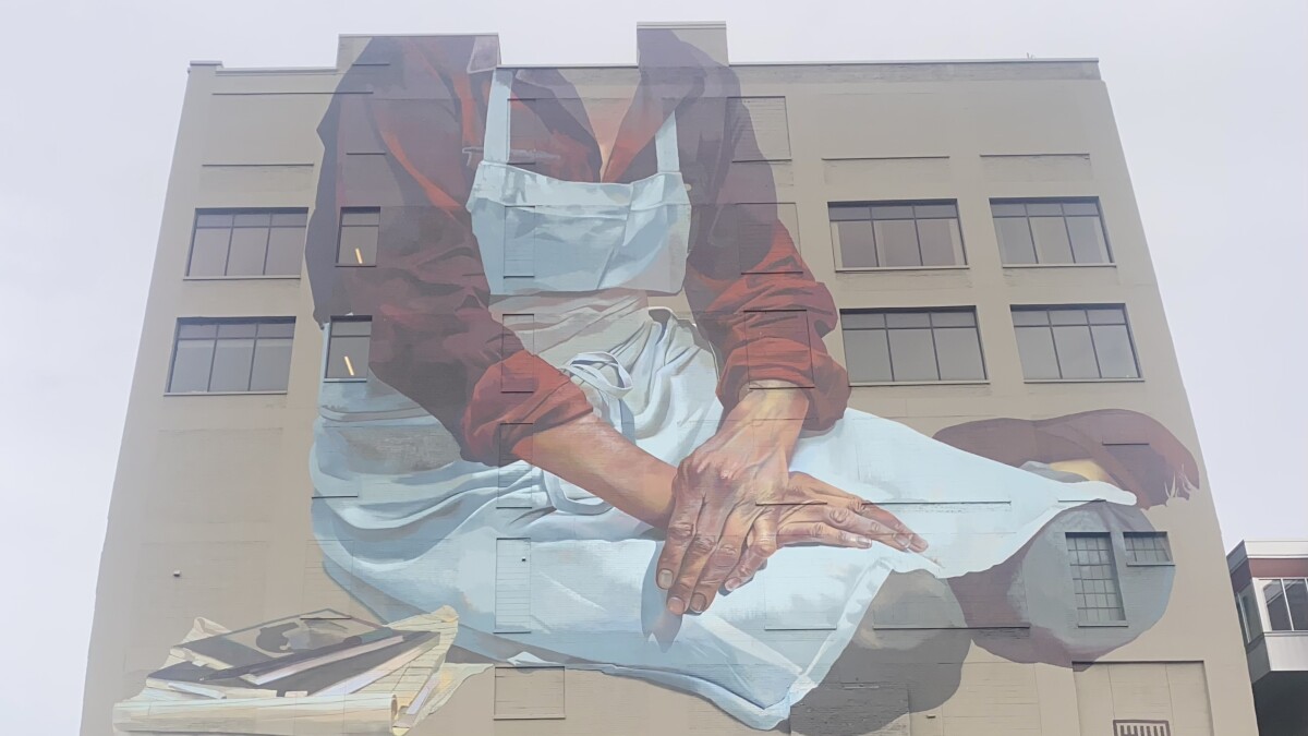 WUWMradio's tweet card. A massive mural in Milwaukee's Third Ward features a seated female figure wearing an apron. Her hands are crossed at the wrist and rest on her knees. But the painting stops at her shoulders. Who is...