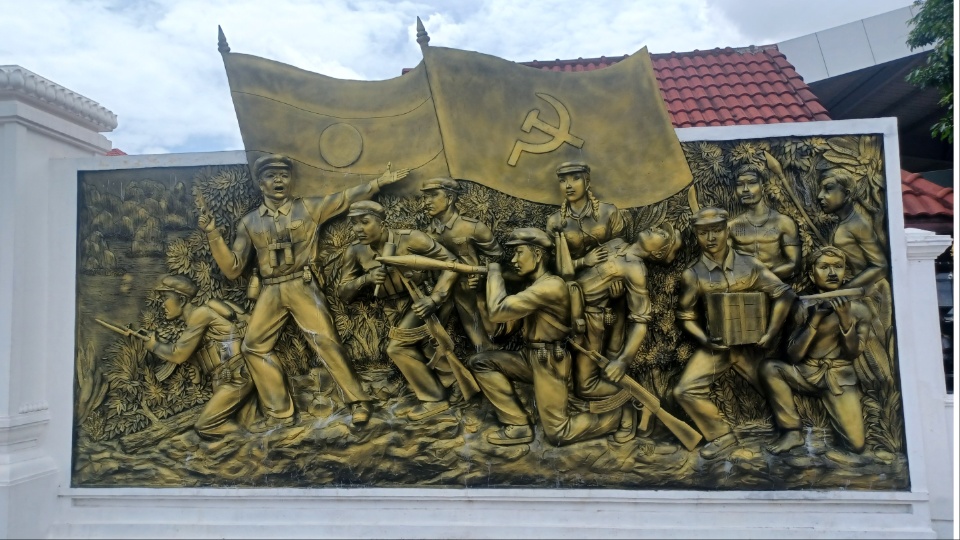 PeoplesWorld's tweet card. VIENTIANE, Laos—Three figures stand atop a raised pedestal, their eyes fixed on the distant horizon: a young woman in traditional dress, raised sickle in hand, and a rifle slung over her shoulder; a...