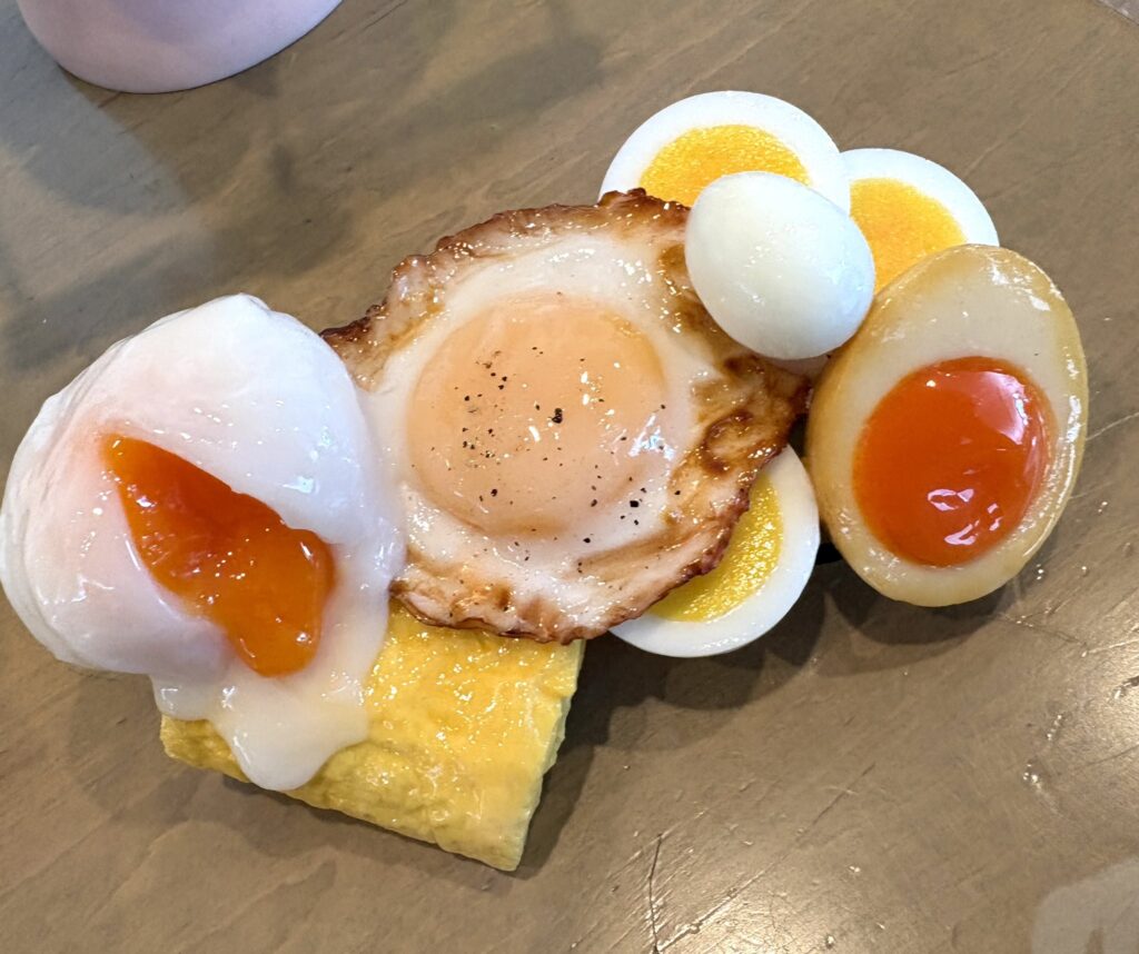 IWASAKI_SAMPLE's tweet card. 株式会社いわさきの食品サンプルがXで、えらい難問をぶつけてきました。投稿では「Q.さて問題です（当てられたらすごい）。温泉卵・目玉焼き・ゆで卵スライス・半熟味玉・卵焼き・うずら玉子…。ありったけの卵を...