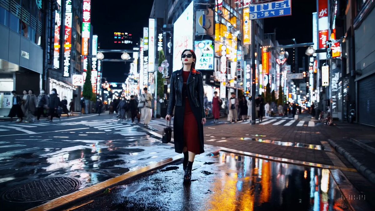 A stylish woman walks down a Tokyo street filled with warm glowing neon and animated city signage. she wears a black leather jacket, a long red dress, and black boots, and carries a black purse.