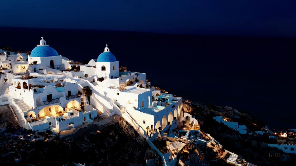 Aerial view of Santorini during the blue hour, showcasing the stunning architecture of white Cycladic buildings with blue domes. The caldera views are breathtaking, and the lighting creates a beautiful, serene atmosphere.