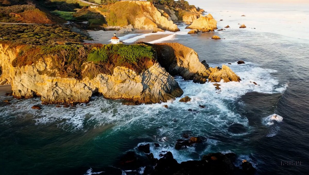 Drone view of waves crashing against the rugged cliffs along Big Sur’s garay point beach. The crashing blue waters create white-tipped waves, while the golden light of the setting sun illuminates the rocky shore.