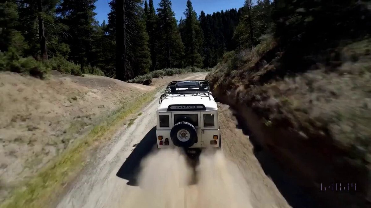 The camera follows behind a white vintage SUV with a black roof rack as it speeds up a steep dirt road surrounded by pine trees on a steep mountain slope, dust kicks up from it’s tires, the sunlight shines on the SUV as it speeds along the dirt road, casting a warm glow over the scene. The dirt road curves gently into the distance, with no other cars or vehicles in sight. The trees on either side of the road are redwoods, with patches of greenery scattered throughout. The car is seen from the rear following the curve with ease, making it seem as if it is on a rugged drive through the rugged terrain. The dirt road itself is surrounded by steep hills and mountains, with a clear blue sky above with wispy clouds.