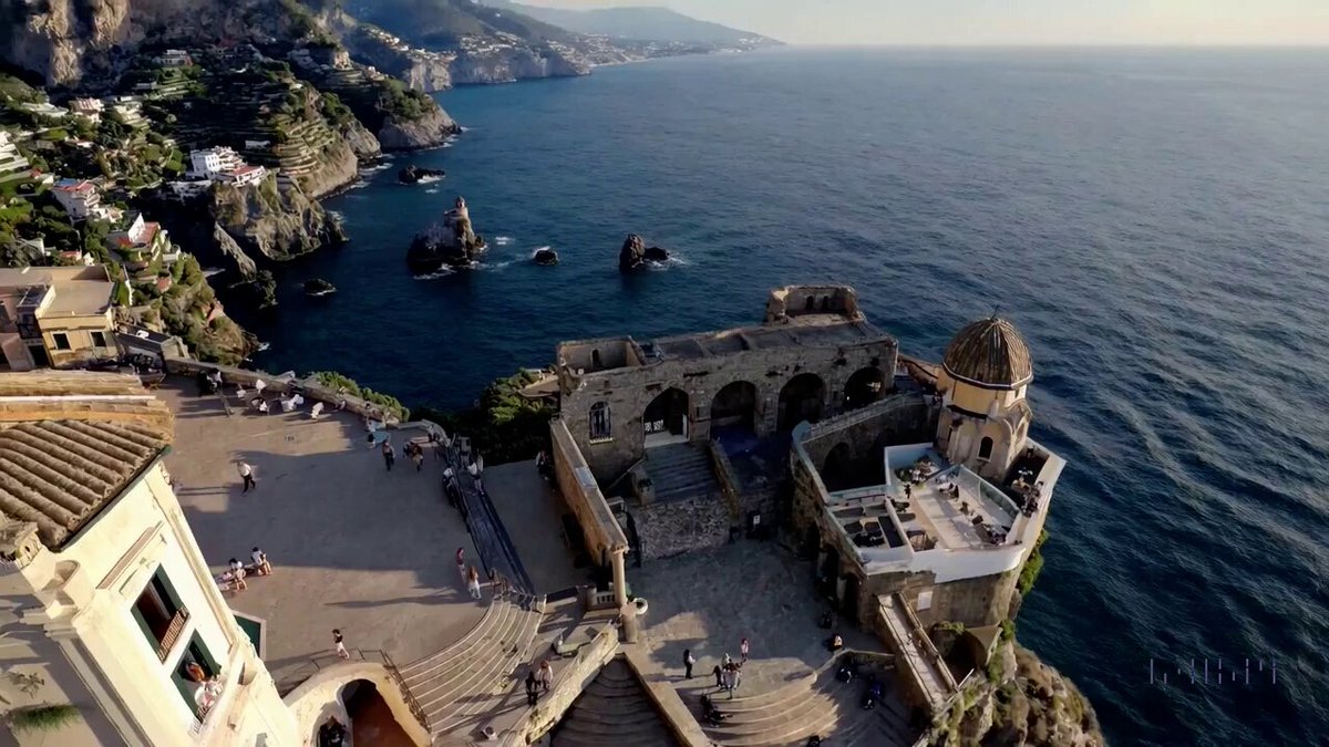 A drone camera circles around a beautiful historic church built on a rocky outcropping along the Amalfi Coast, the view showcases historic and magnificent architectural details and tiered pathways and patios, waves are seen crashing against the rocks below as the view overlooks the horizon of the coastal waters and hilly landscapes of the Amalfi Coast Italy, several distant people are seen walking and enjoying vistas on patios of the dramatic ocean views, the warm glow of the afternoon sun creates a magical and romantic feeling to the scene, the view is stunning captured with beautiful photography.
