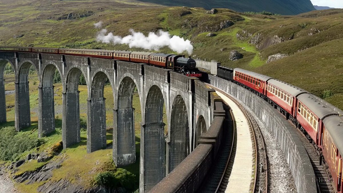 The Glenfinnan Viaduct is a historic railway bridge in Scotland, UK, that crosses over the west highland line between the towns of Mallaig and Fort William. It is a stunning sight as a steam train leaves the bridge, traveling over the arch-covered viaduct. The landscape is dotted with lush greenery and rocky mountains, creating a picturesque backdrop for the train journey. The sky is blue and the sun is shining, making for a beautiful day to explore this majestic spot.