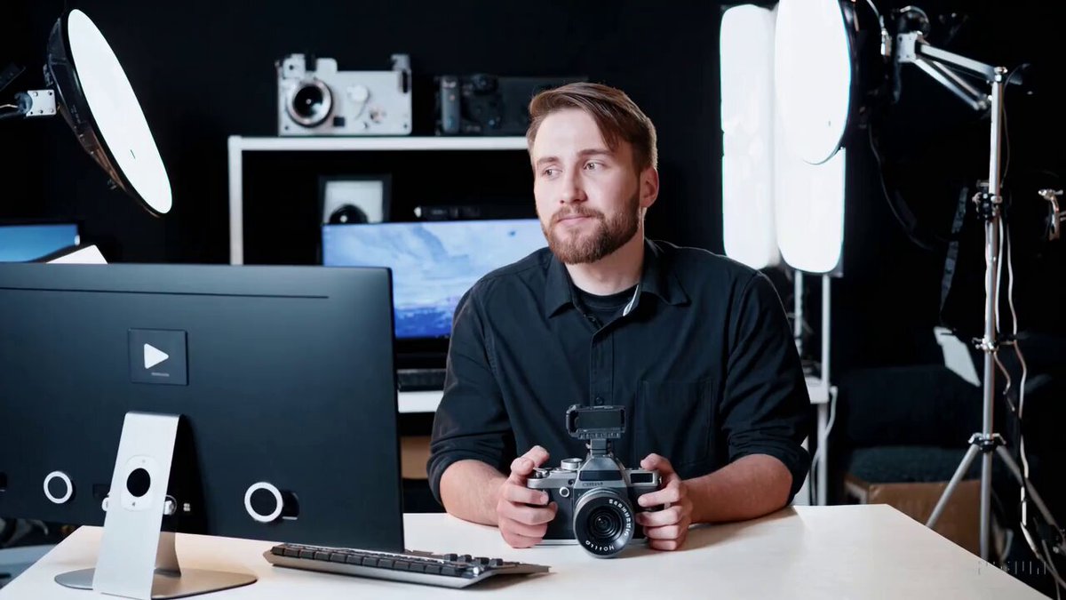 A young professional product reviewer in a well lit video studio is surrounded by gadgets and technology, sitting in front of a computer with two displays. He's holding a cinema camera as he ponders what video to make next. He is in focus, while the background is slightly blurred for cinematic effect