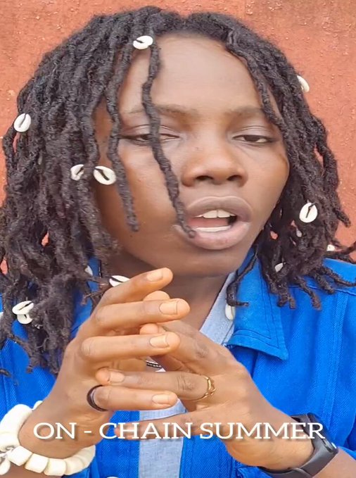 Person with dreadlocks adorned with shells, wearing a blue shirt, gesturing with hands, with 'ON - CHAIN SUMMER' text overlay
