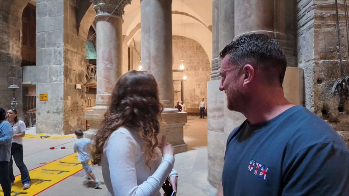 The image shows the interior of an ancient stone church with tall columns, arches, and hanging lamps. In the foreground, a man with short gray hair, beard, and glasses, wearing a blue t-shirt with a UK flag patch, is speaking to a young woman with long wavy brown hair in a white sweater. In the background, other people including a child in white clothing and adults in casual attire are visible near yellow caution tape on the floor and red markers, suggesting ongoing maintenance or restricted areas.