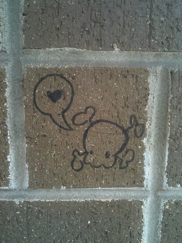 Drawn on a brick wall in black permanent marker: a small skull-and-crossbones has a word balloon with a heart in it