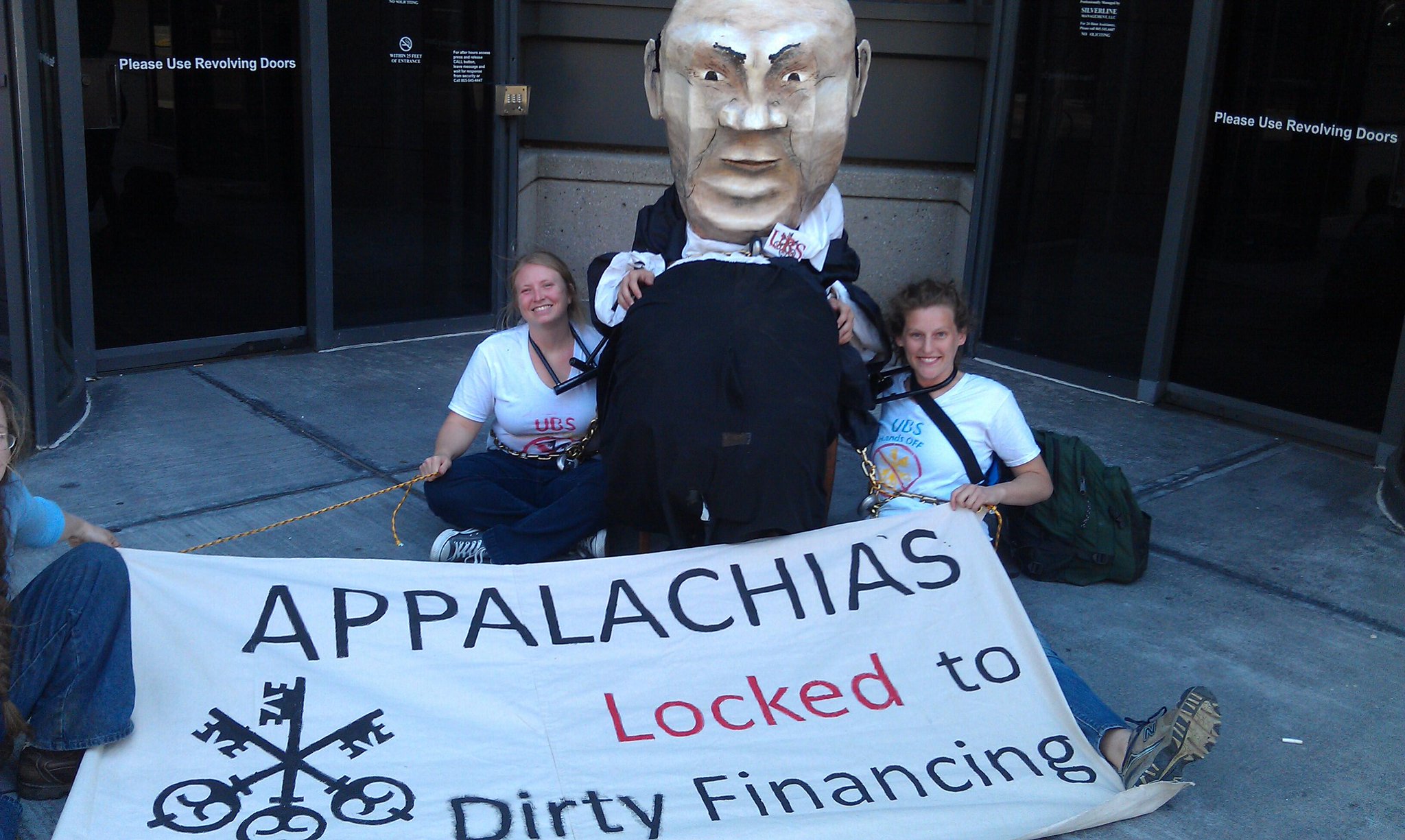 Two women locked to fat cat banker puppet at UBS Knoxville office.