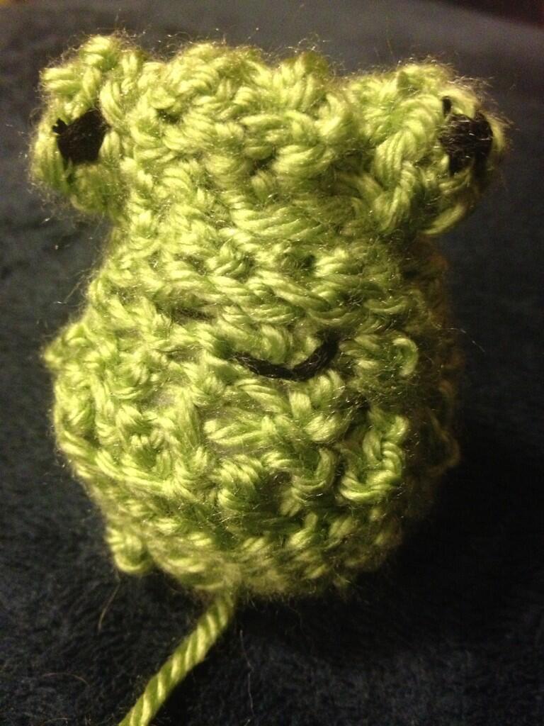 Head of a crocheted frog with embroidered black eyes and smile. Still working on the extra body parts