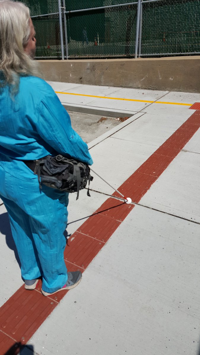 A Pedestrian With Long White Hair And A Teal Outfit Uses A White Cane On A A Pedestrian With Long White Hair And A Teal Outfit Uses A White Cane On A