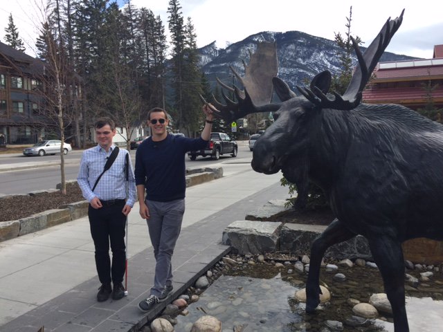 Grant Hardy and Anthony McLachlan stand beside a giant moose statue with tall trees and snow capped mountains in the background.