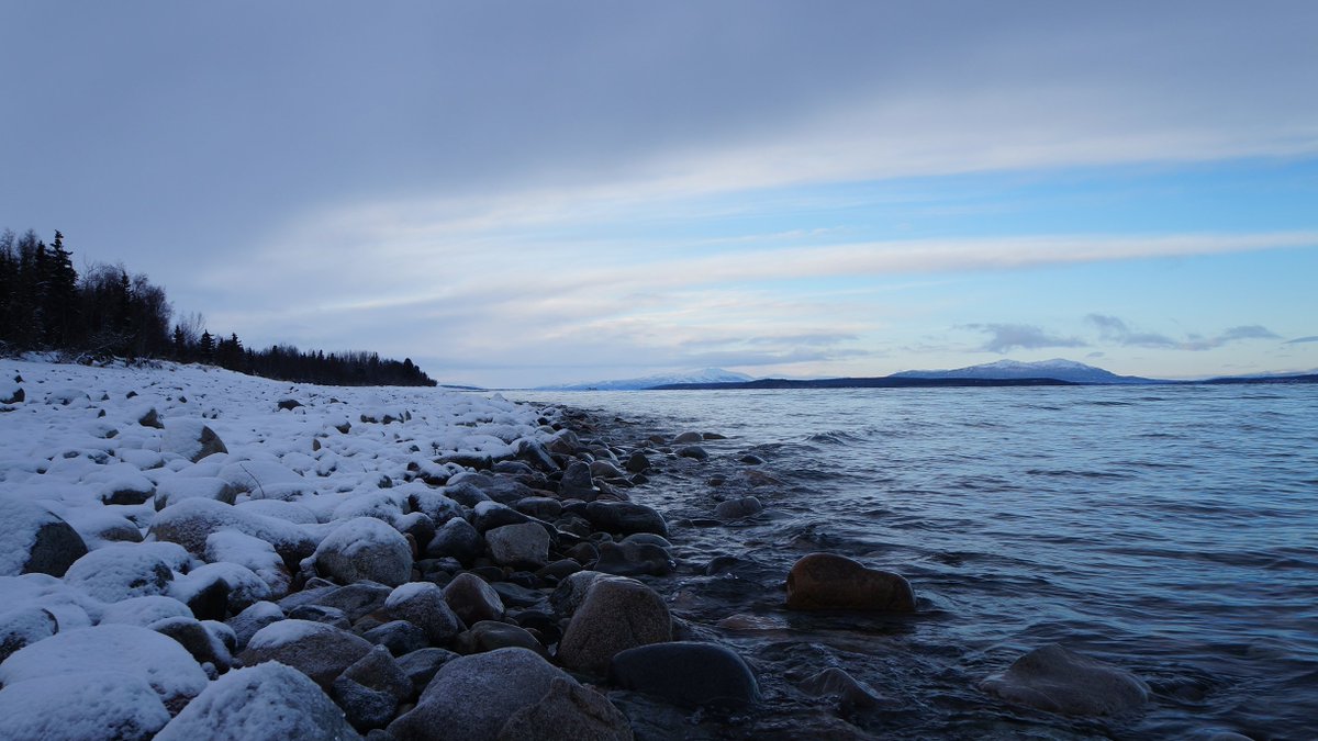 A rocky beach on the lake shore with snow covered rocks, low clouds, and dim skies.