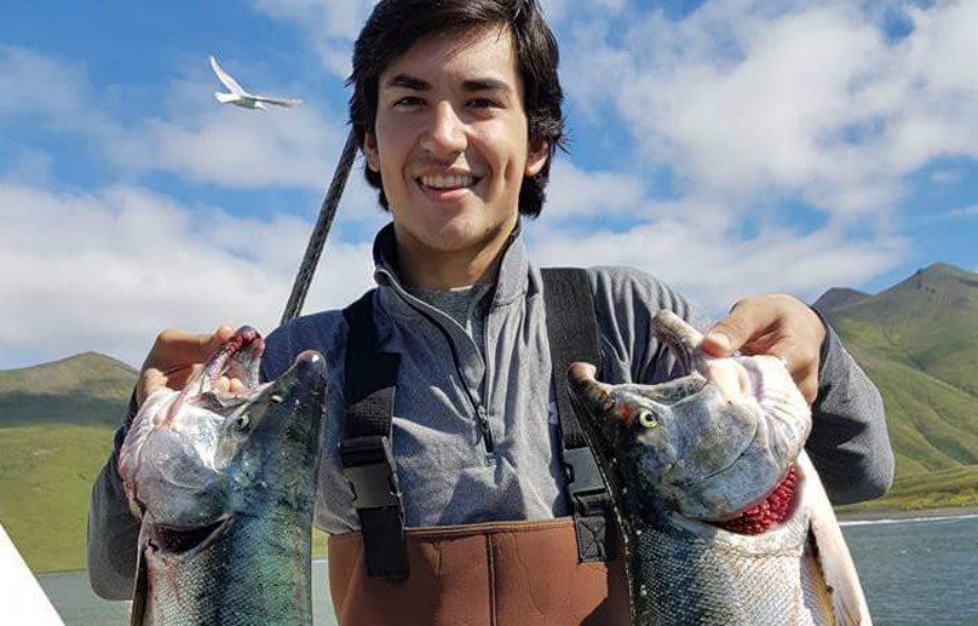 A young man stands on a boat holding up two large fish.