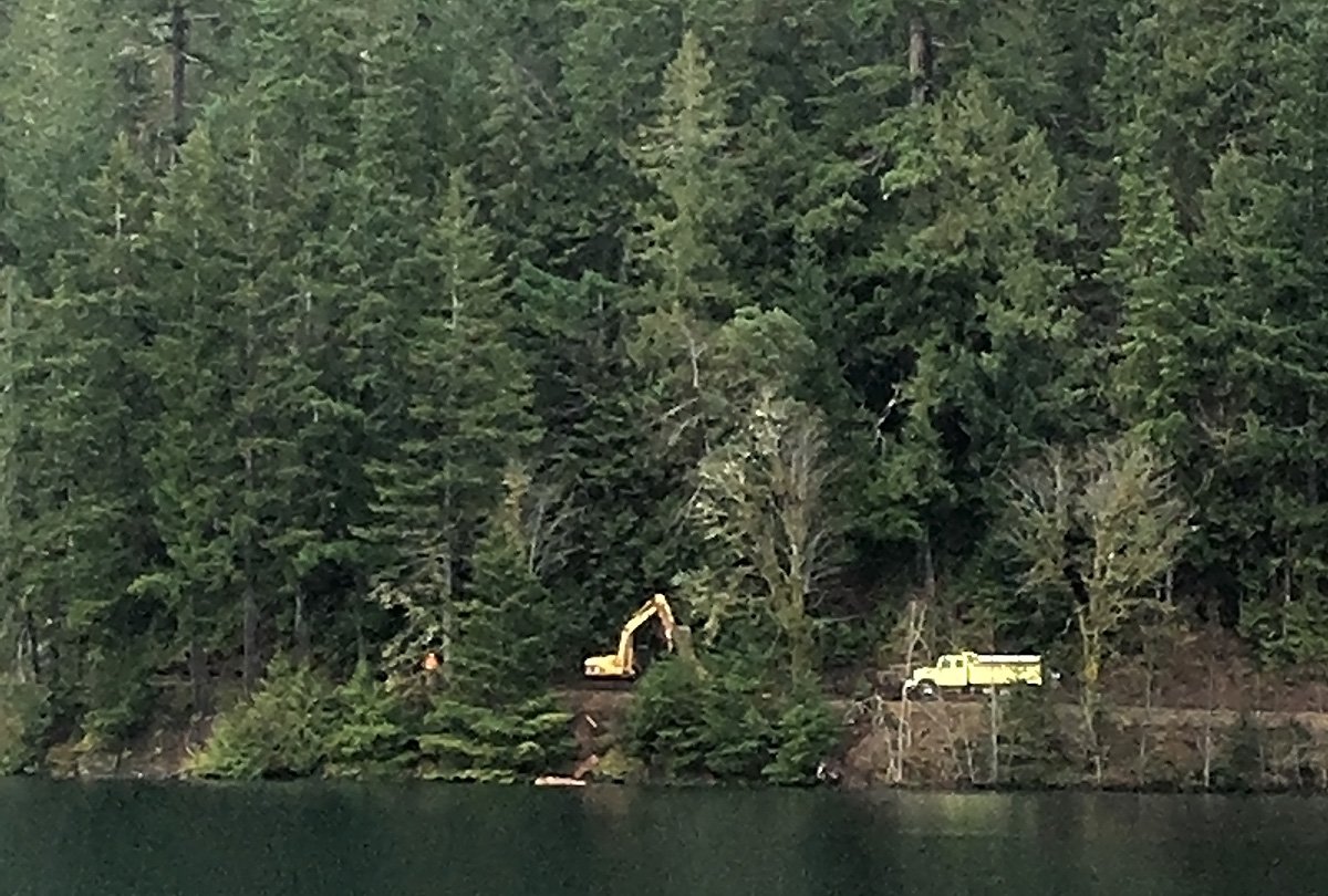 yellow dump truck and a yellow excavator on a roadway next to a lake, many evergreen trees