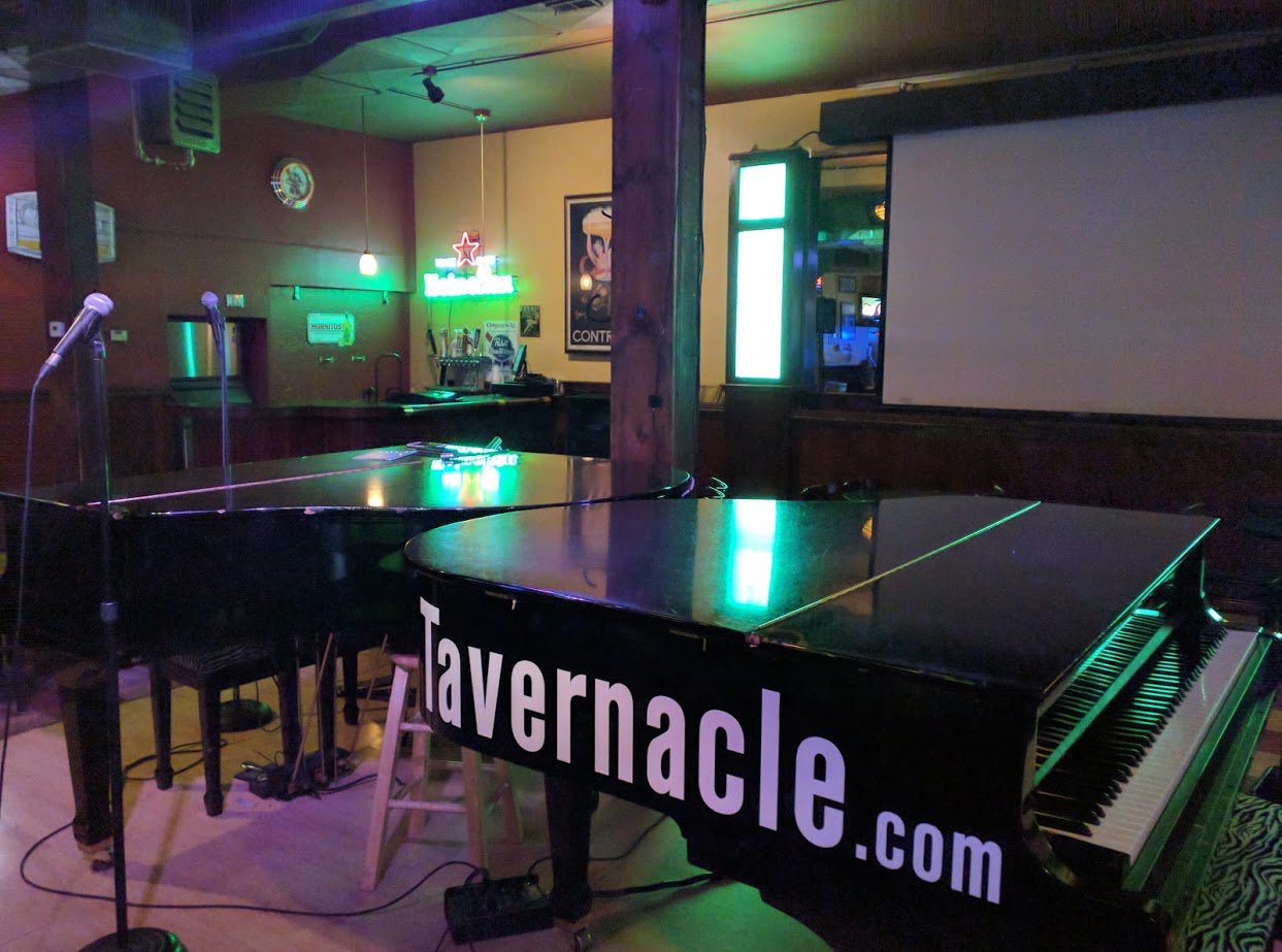 2 dueling pianos that have Tavernacle stenciled onto them. In a bar with a mic on a stage.
