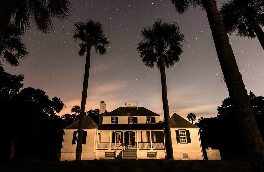 A large white plantation house with palm trees in its front yard stands under a night sky.