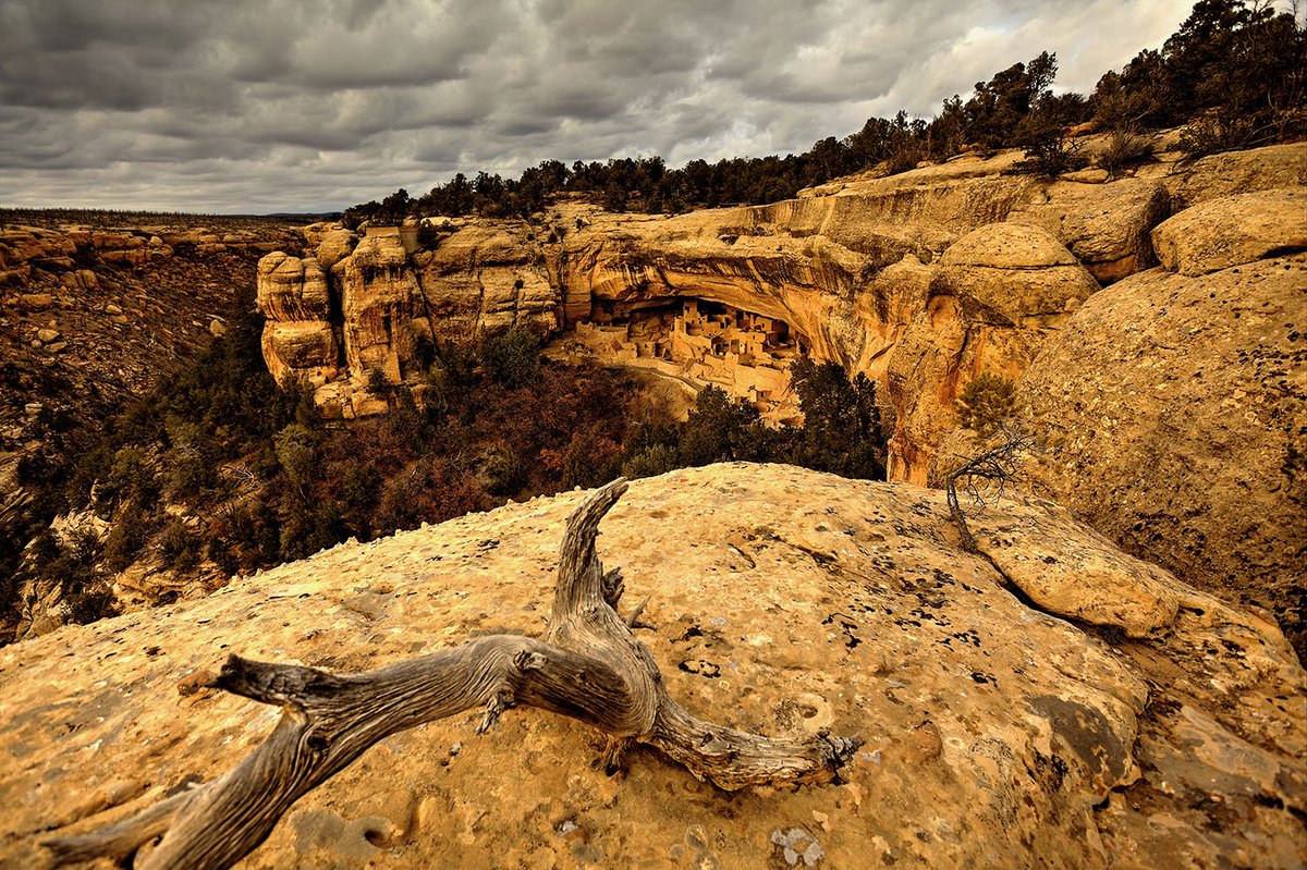 A massive canyon wall curves across a desert landscape under a cloudy sky. Under a large overhang of the cliff sits a small village of brick buildings.