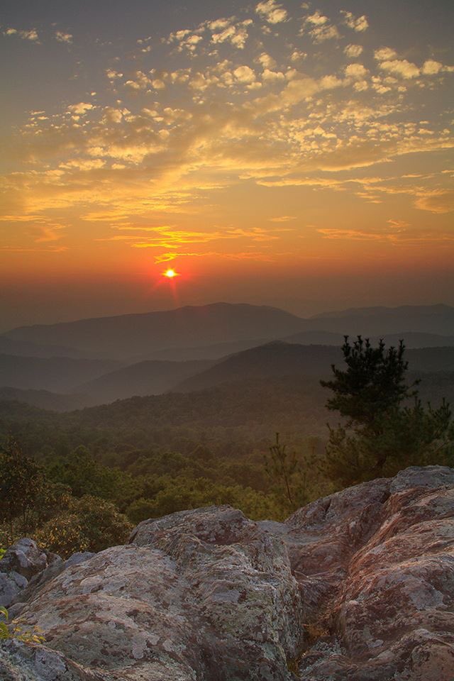 A orange sun appears over rolling hills as seen from a rocky overlook