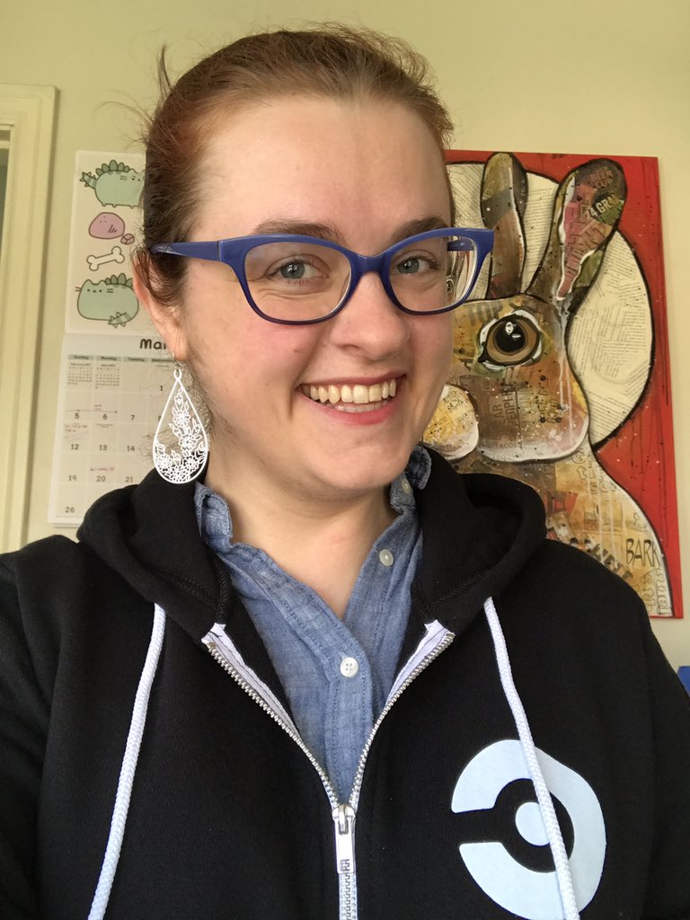 A smiling woman (me!) wearing a black CircleCI logo hoodie. In the background, a Pusheen calendar (March) and a collage painting of a brown bunny in front of a full moon on a red background.