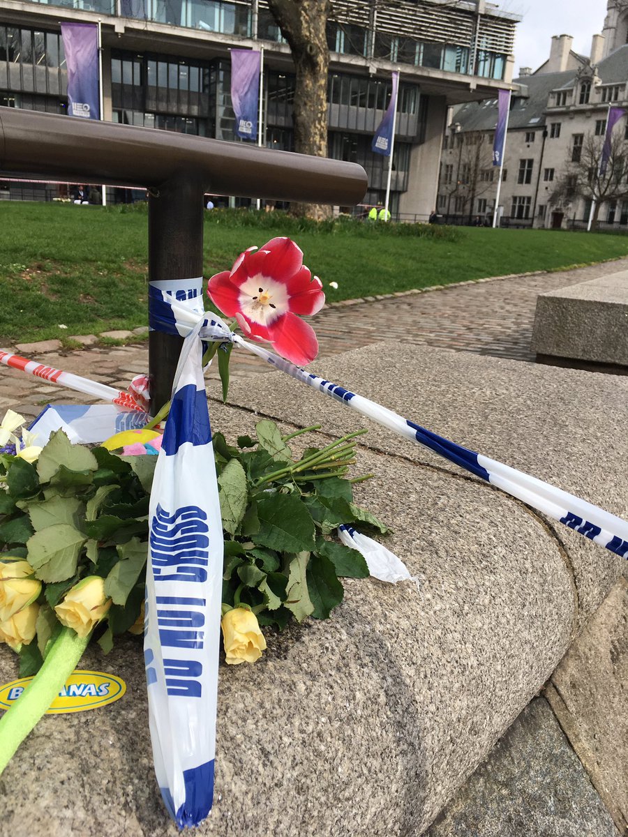 A railing, overlooking the QEII conference centre, with flowers tucked into a "Police line - DO NOT CROSS" tape