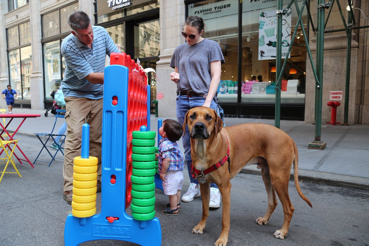 A family stands beside a super-sized game of connect-four while standing in a street that is closed to vehicles and open to the public. A folding table and folding chair is in the street behind the family. The family dog is looking at the photographer.