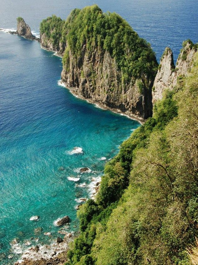 A thin, rocky outcropping covered in trees juts out into the clear, blue water of the Pacific Ocean from a tree covered shoreline in American Samoa.