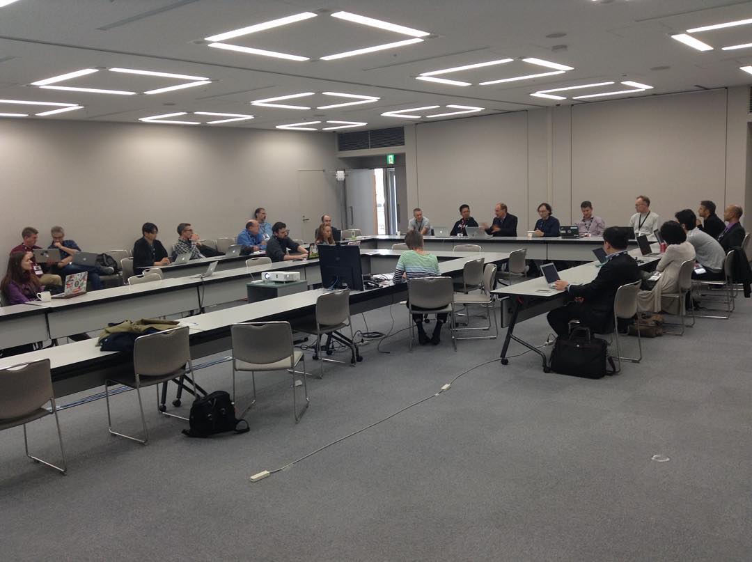 a group of people sitting around a set of conference tables arrange in a U-formation, inside a large meeting room.