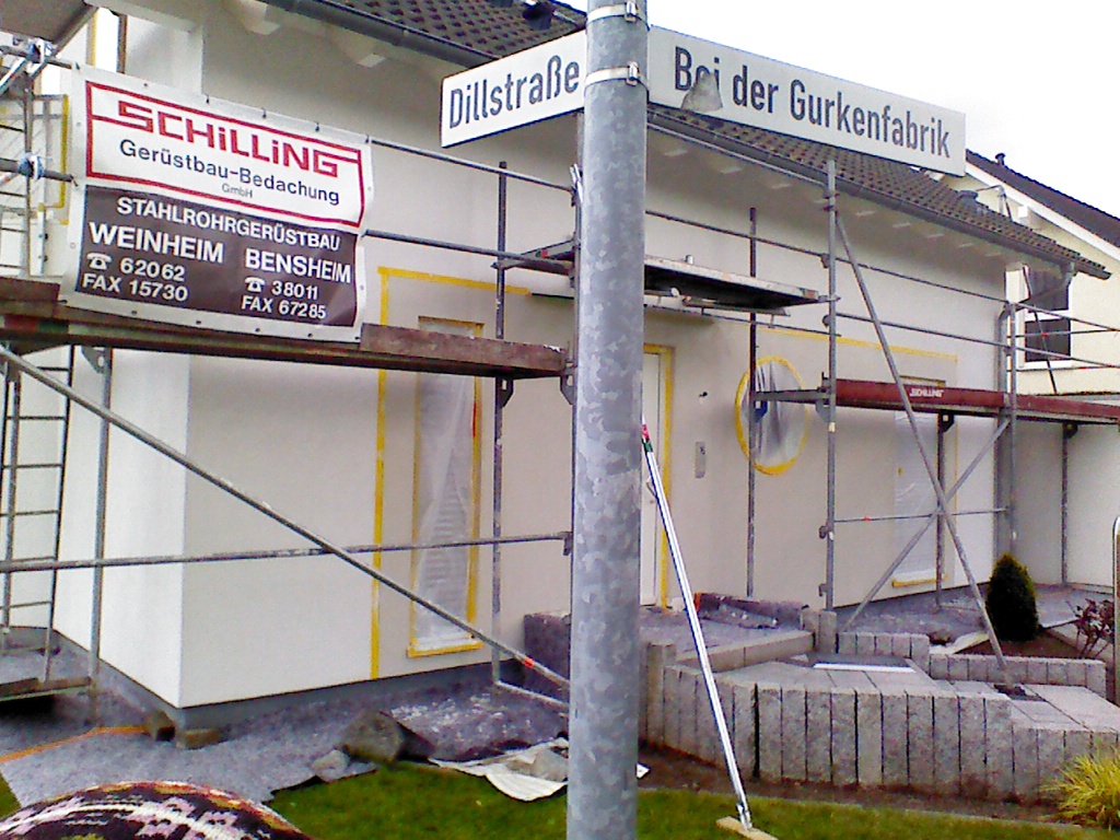 Ein Neubau mit Gerüst. Davor ein Pfosten mit zwei Straßenschilder: Links steht "Dillstraße" und rechts "Bei der Gurkenfabrik".