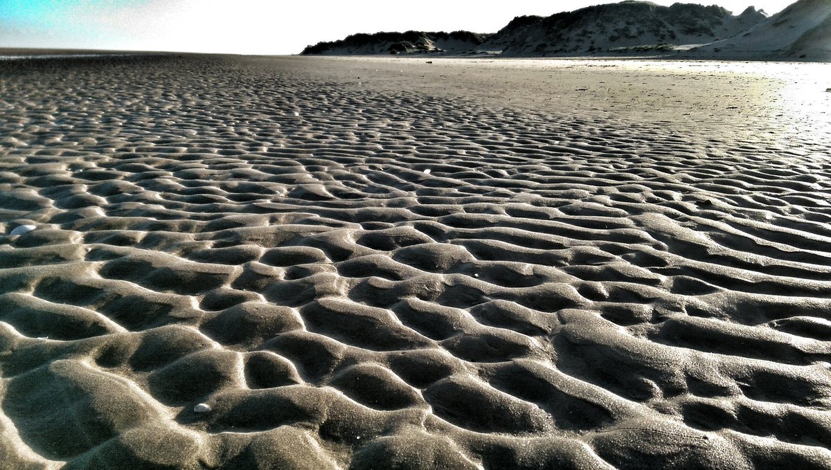 Sun casting shadows through the ripples in the sand caused by the coastal tide; with sand dunes in the background.