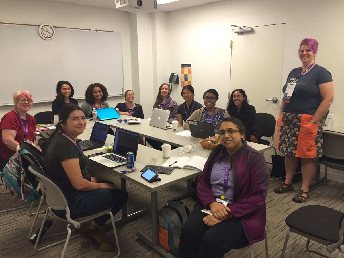 A group of 10 women sitting around a cluster of tables. They have laptops in front of them and have turned to smile at the camera.