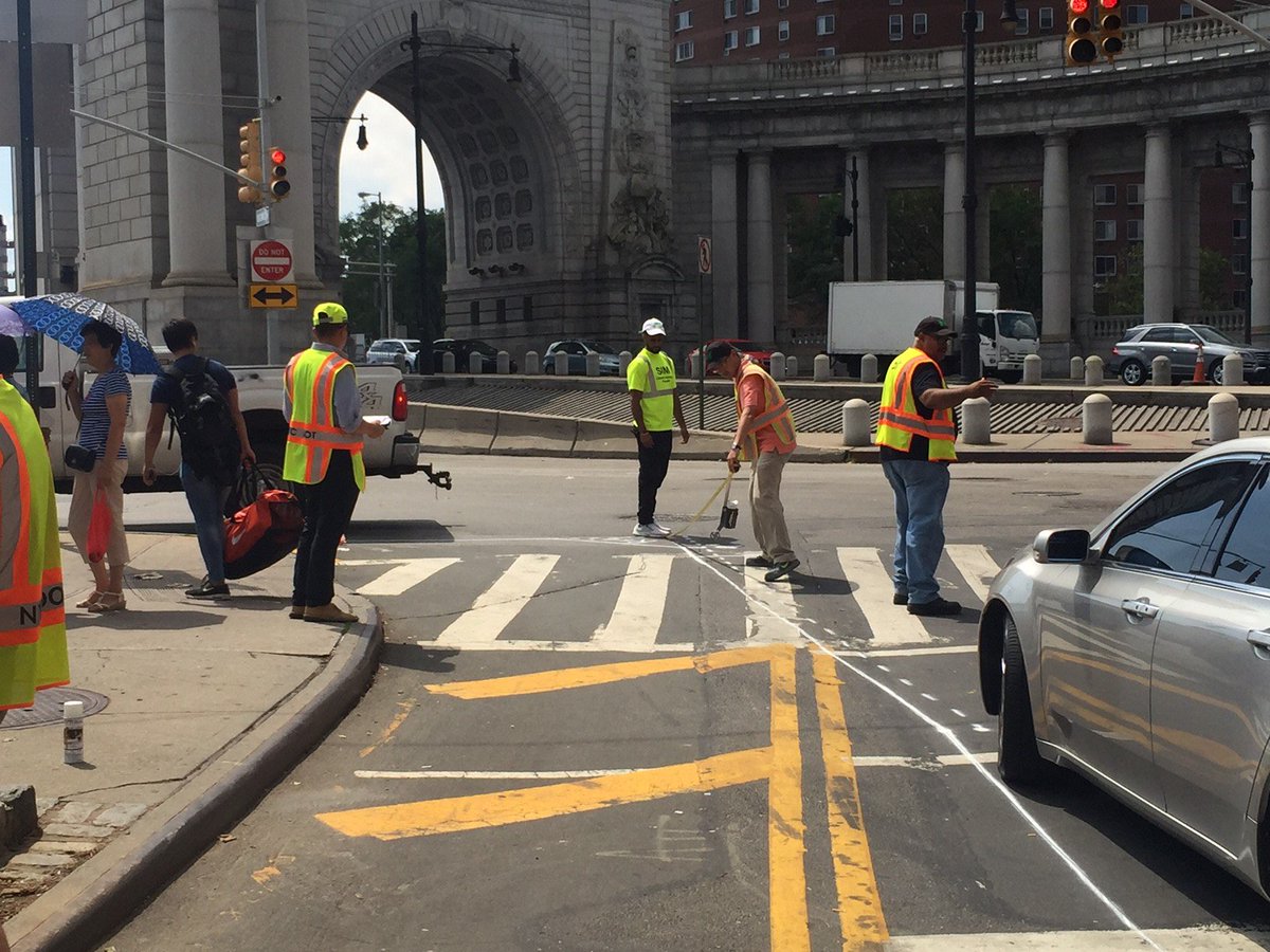 Crews in bright safety vests spray paint on the roadway where new concrete island will be install. Pedestrians stand on the current pedestrian median. The Manhattan Bridge arch is visible int he background.
