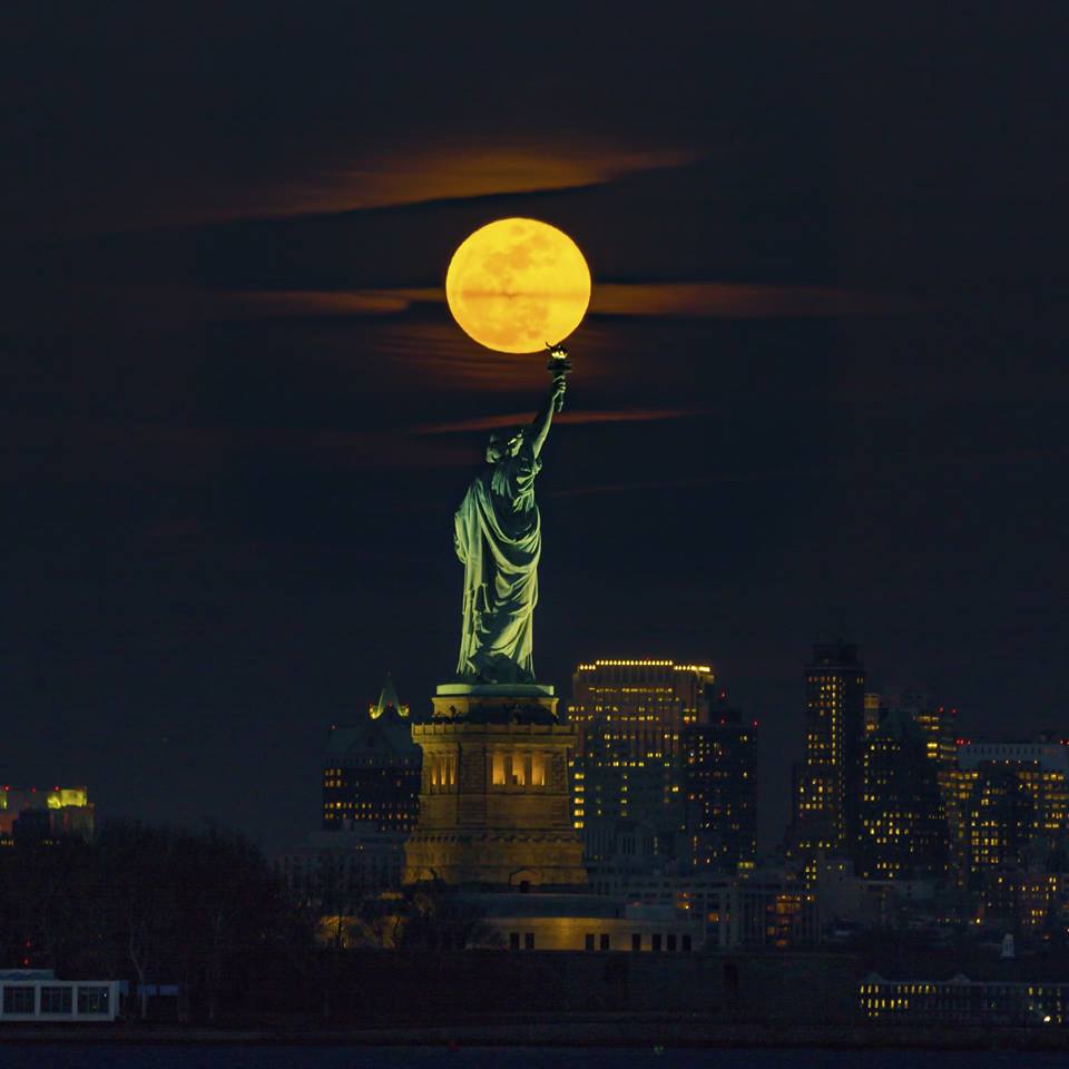 The statue of liberty stands on a tall pedestal holding a torch up high with buildings of New York City in the background and a huge full moon shining above it.