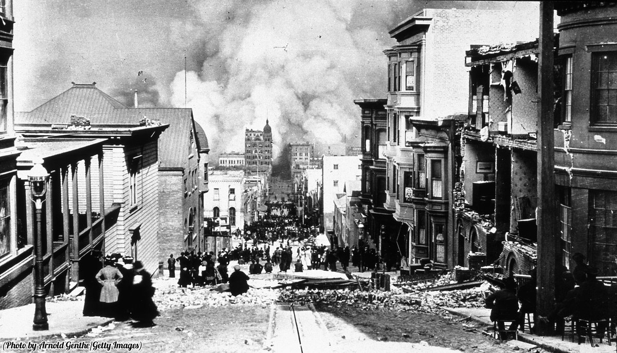 Crumbling buildings line a street and smoke rises in the background after the San Francisco earthquake, April, 1906.
