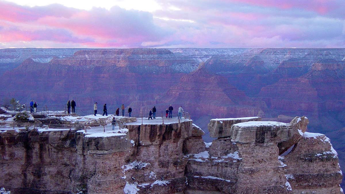 tiny people wearing winter jackets looking out behind a guardrail from a scenic overlook at pink sunset clouds and snow capped peaks..