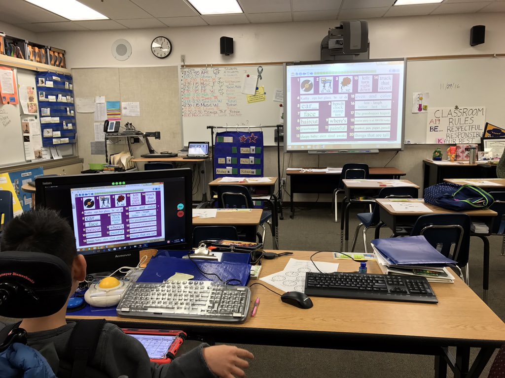 Photo from back of the room shows online learning activity on SmartBoard and student seated in back row with same activity on desktop monitor