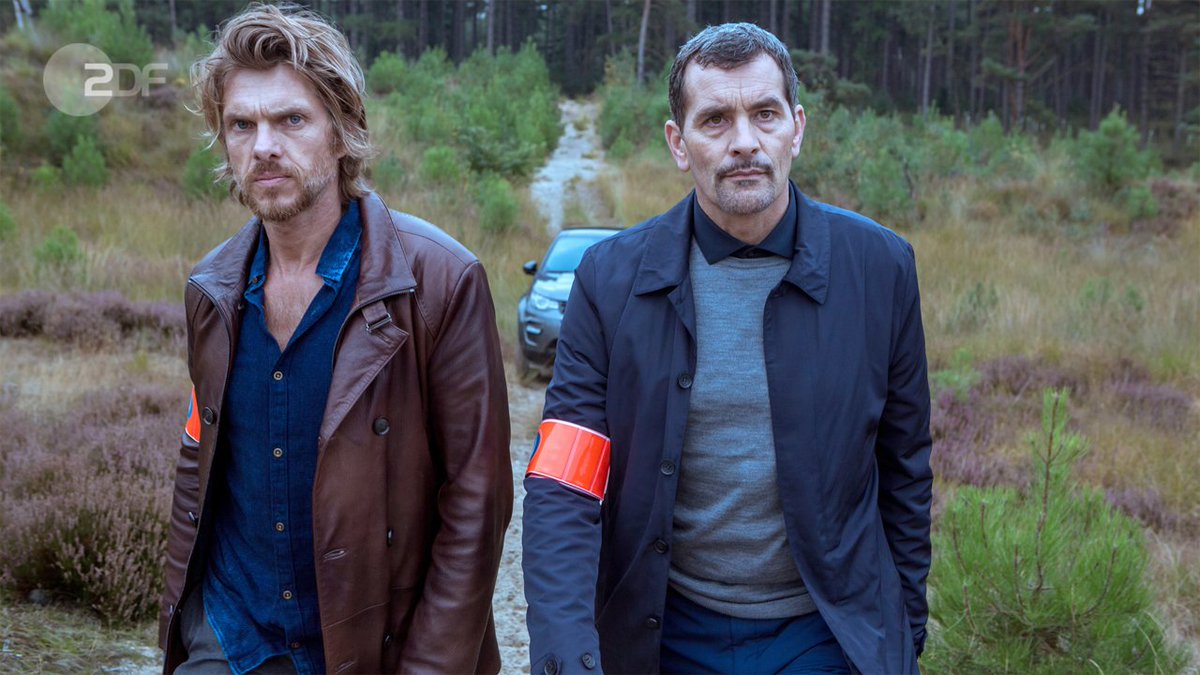 "Das letzte Opfer": Verstuyft (Werner De Smedt) und Vincke (Koen De Vouw) laufen über einen Feldweg auf die Kamera zu. Beide tragen eine rote Armbinde am rechten Arm. Im Hintergrund steht ihr Wagen auf einem Waldweg.